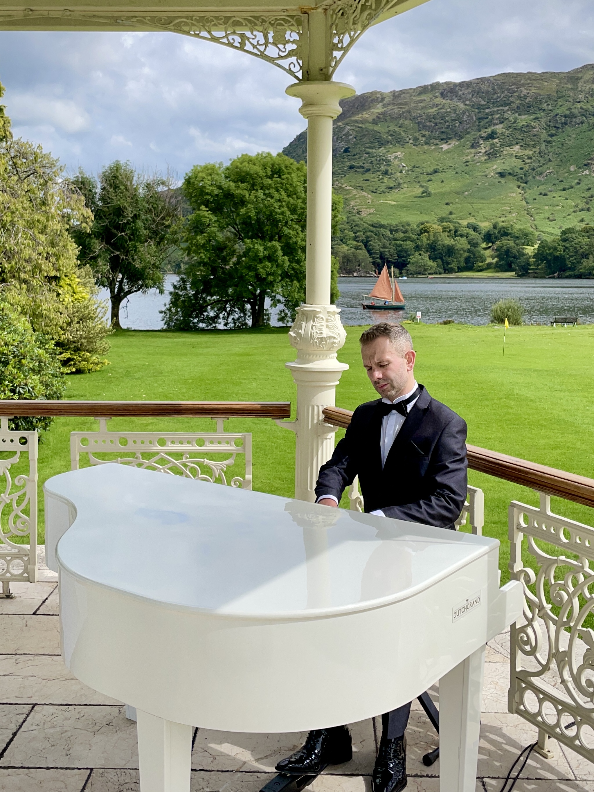 Wedding pianist Craig Smith performs, dressed in a tuxedo, on a white piano in the lakeside gazebo at the Inn on the Lake, with an orange sail boat on Ullswater behind him and mountains in the background